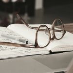 A pair of glasses resting on an open notebook filled with handwritten notes beside a laptop.