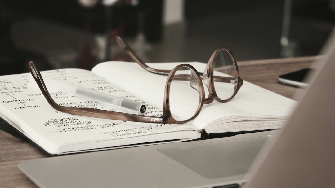A pair of glasses resting on an open notebook filled with handwritten notes beside a laptop.